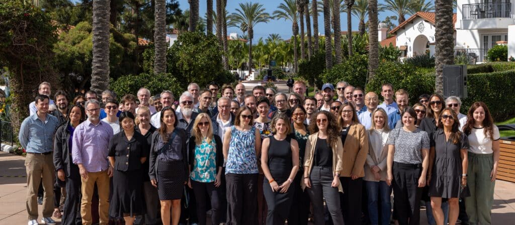 Photo of about 80 participants at the openRxiv Day. People are gathered together facing forward toward the camera and smiling. It's sunny and in the background are palm trees and white buildings with tile red roofs.