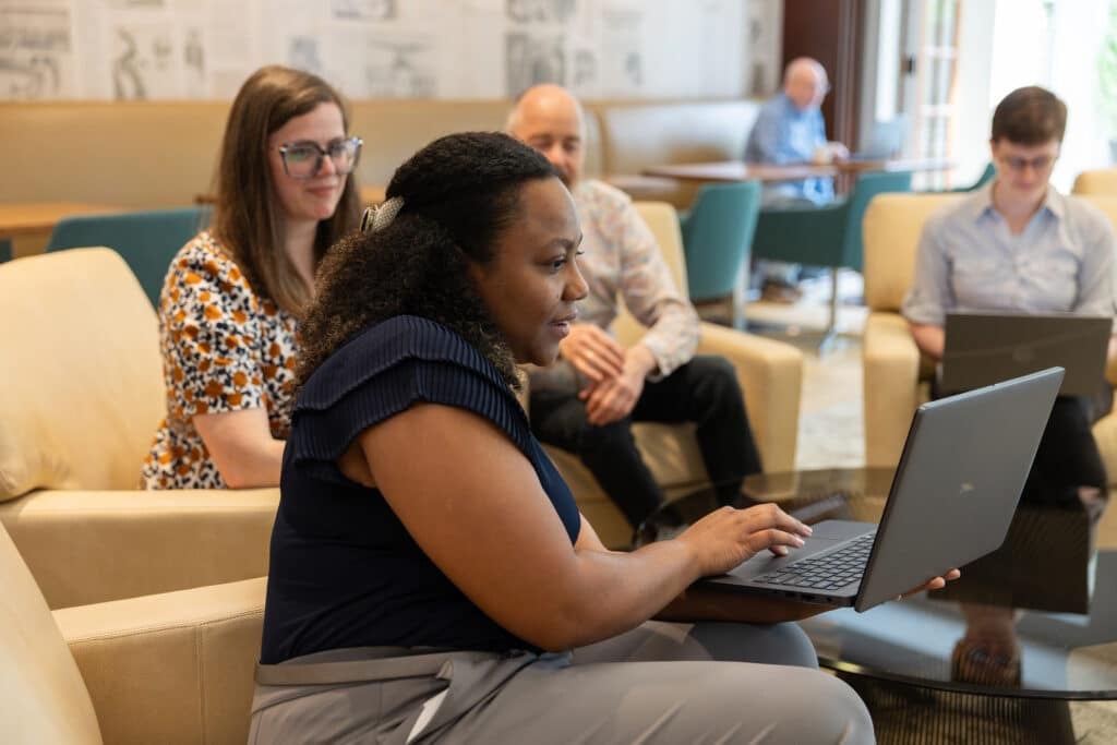 Four people sitting on comfortable  yellow chairs around a coffee table looking at what a woman is showing them on a laptop and smiling. 